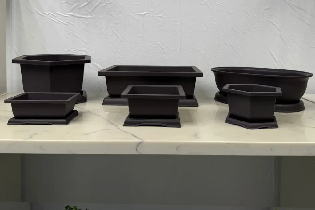 A selection of plastic bonsai training pots of different sizes, all with trays beneath, on an indoor marble